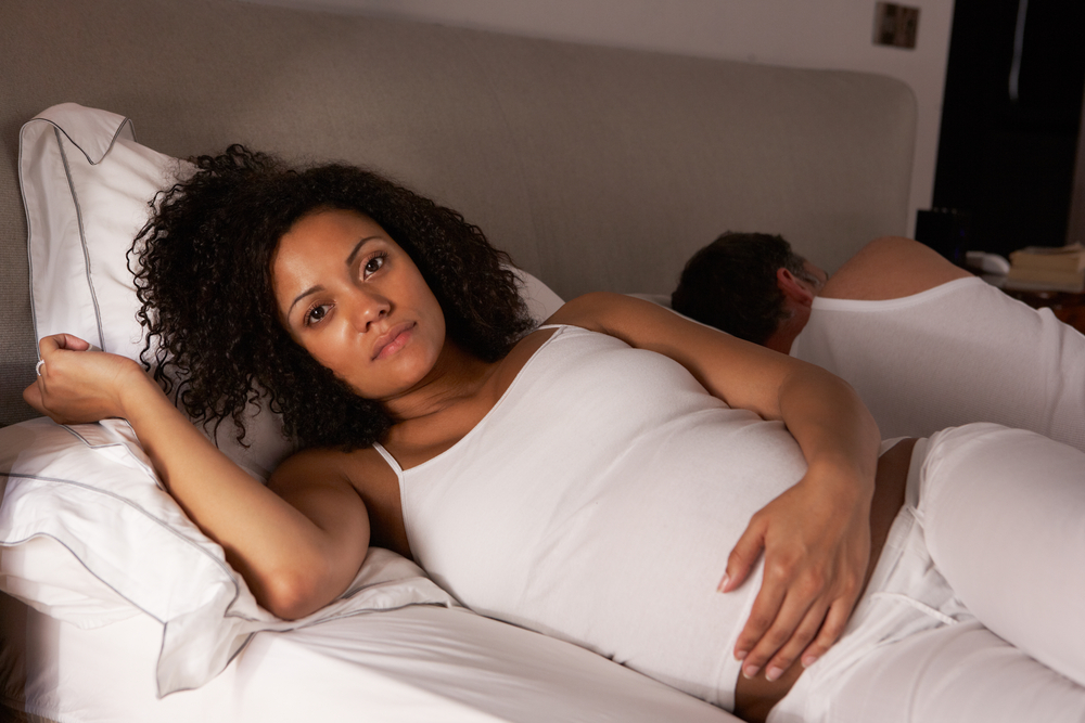 A pregnant woman lays in bed with her eyes open, her partner asleep next to her.