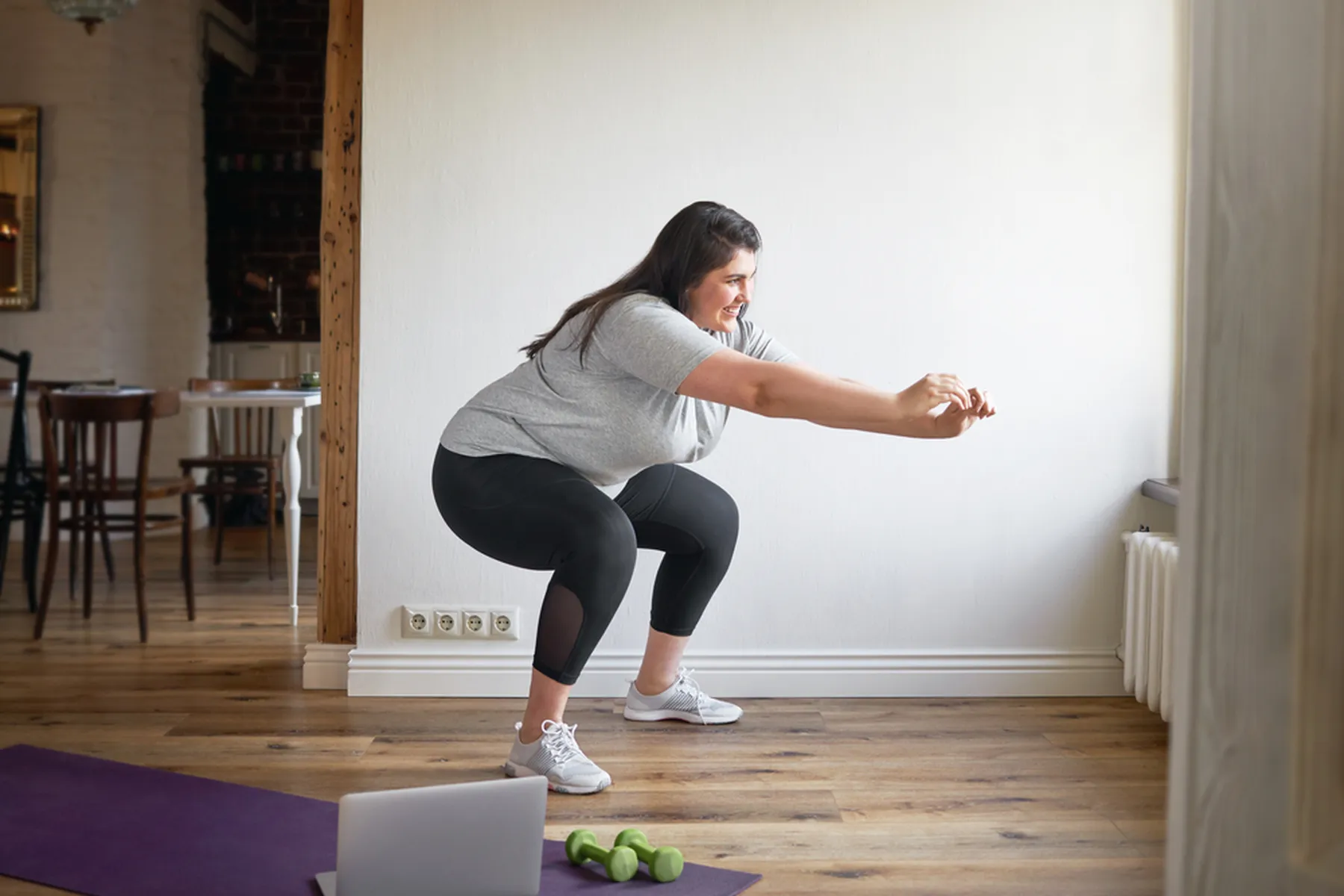 A woman in a grey t-shirt and black workout pants does a squat as she straightens her arms out in front of her. 