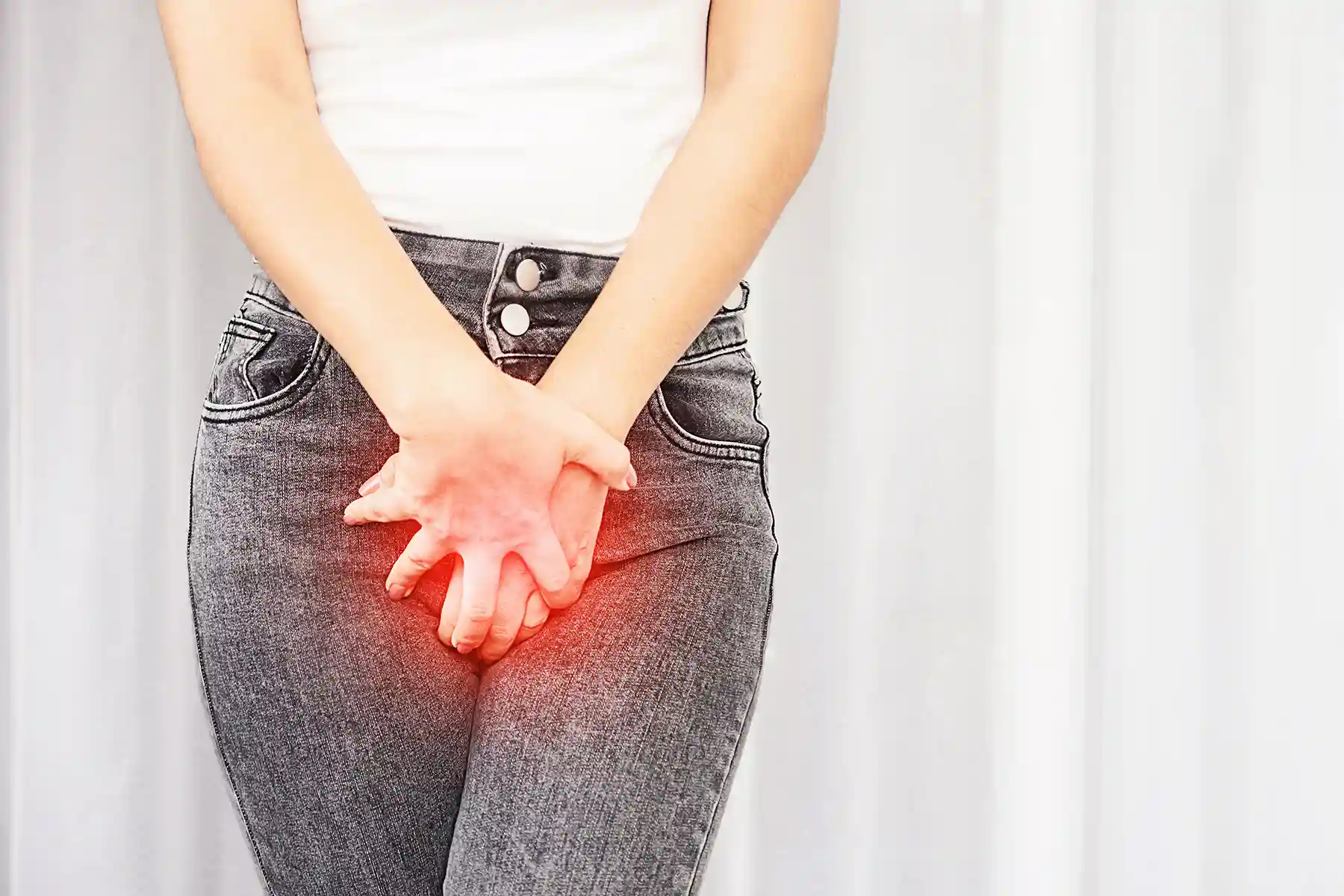 A woman in a a white shirt and black stonewashed jeans holds her hands to her genital region. There is a red glowing color under her hands to signify pain or burning. 