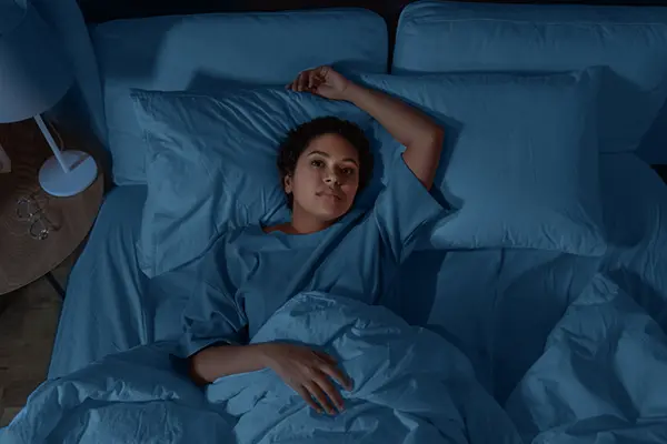 A woman lying on her bed in bed in a dark room. One hand rests on her stomach while the other is above her head on a pillow. 