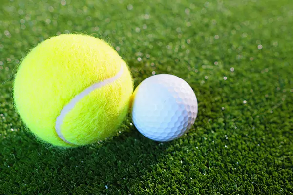 A tennis ball and a golf ball sitting next to each other on green turf or grass. 