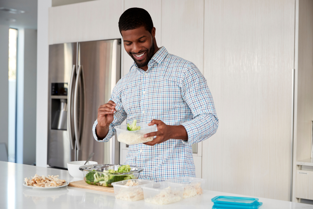 A man stands in his kitchen, smiling, and meal preps with healthy foods.