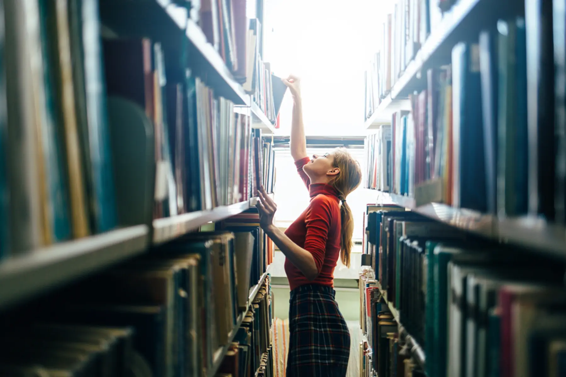 A librarian stand in the stacks and reaches up to grab a book.