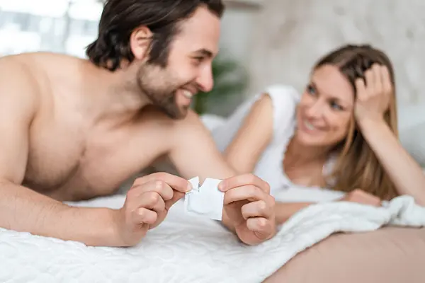 A couple lays in bed and the man holds a condom.