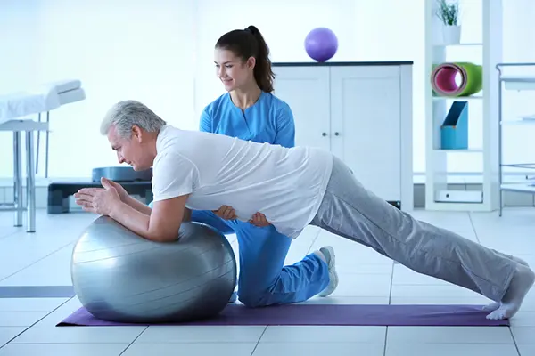 A man at a session with a physical therapist. He is doing a plank while supporting his arms on a large ball. The physical therapist has her hands under his torso for extra support. 