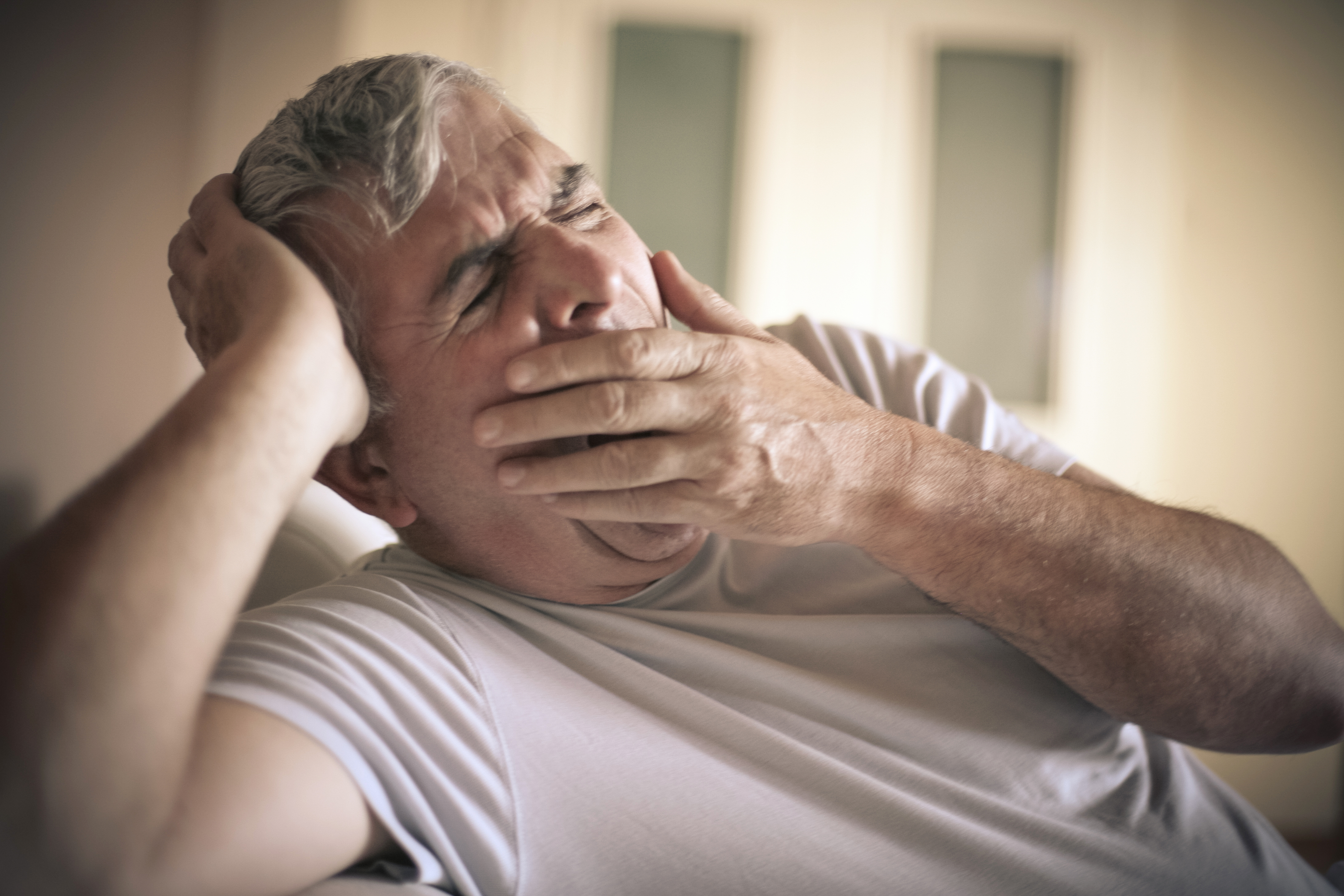 An older man in a light-colored t-shirt lets out a big yawn as he covers his mouth with his left hand. His eyes are closed, and his head is resting on his right hand. 
