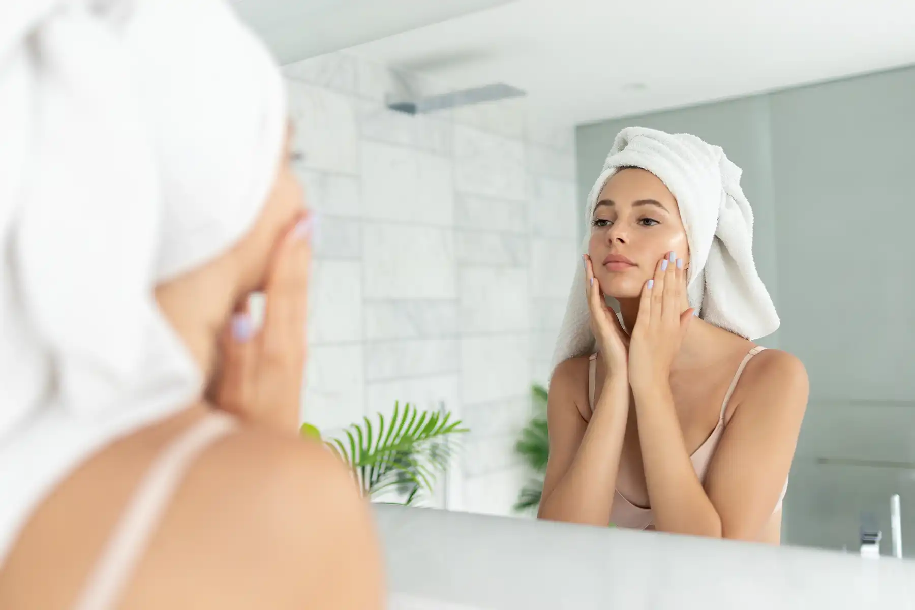 Woman wearing a towel on her head applying moisturizer to her face and looking into the mirror.