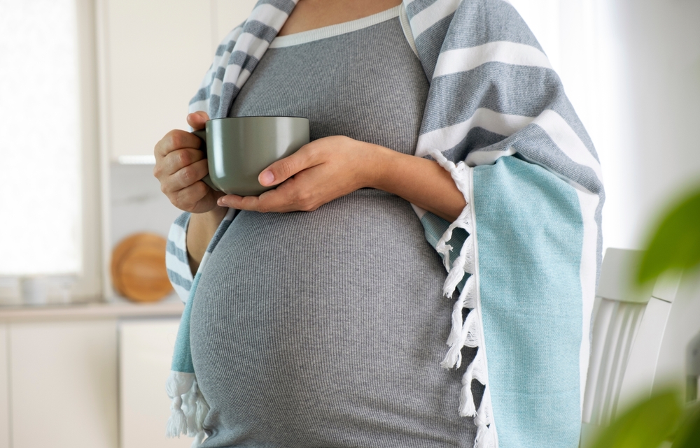 A pregnant woman stands with a blanket over her shoulders and holds a cup of tea.