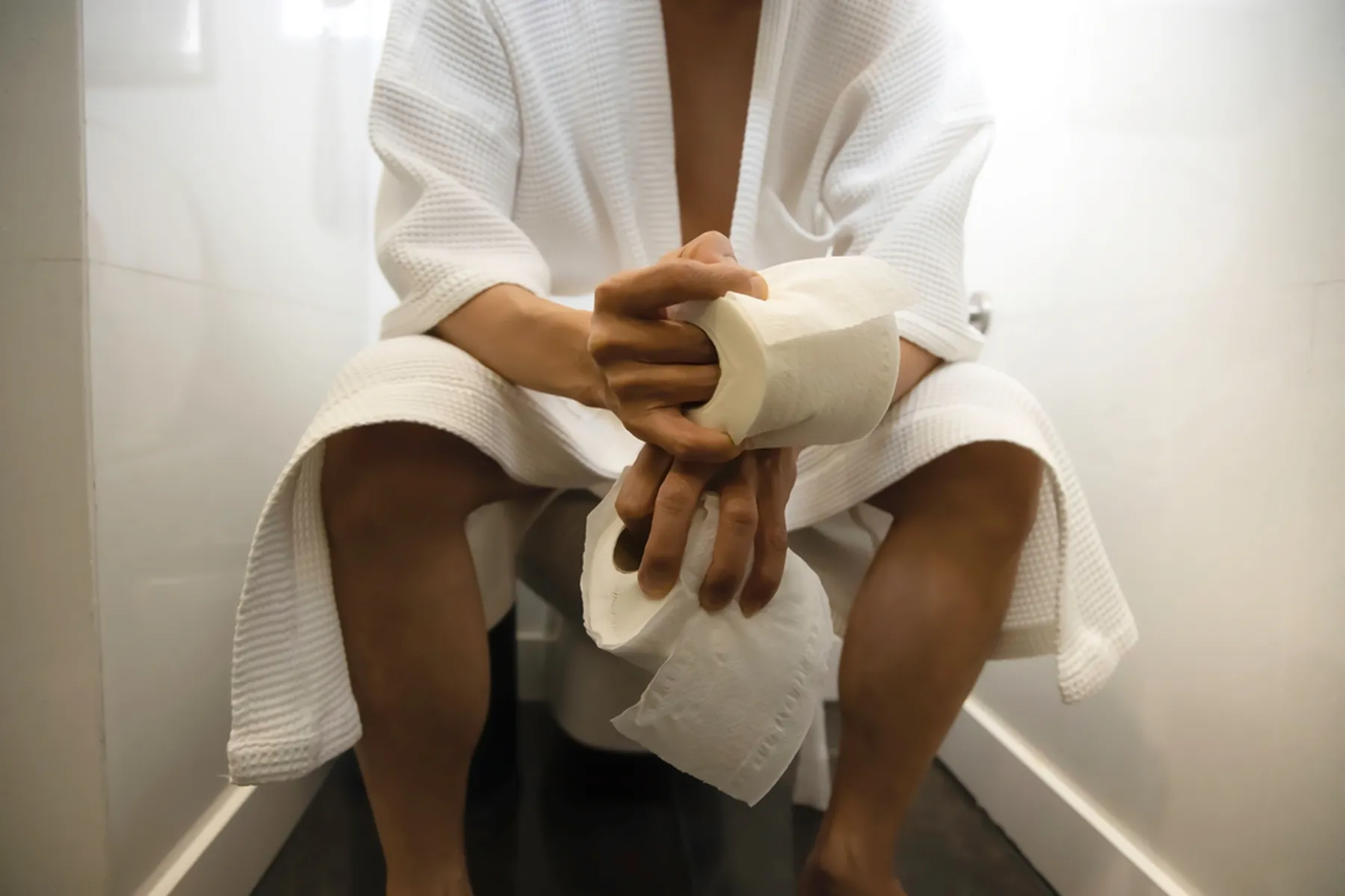 Man sitting on toilet in robe holding toilet paper.