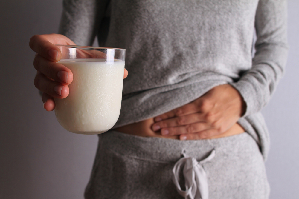A woman holds a glass of milk next to her stomach, which she clutches with her other hand.