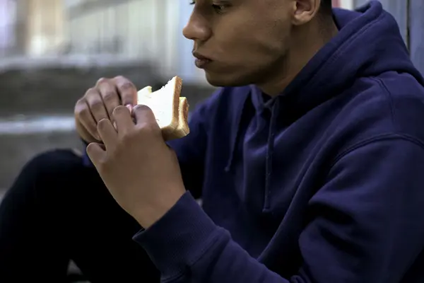 A man sits and makes a sad face while biting into a sandwich.