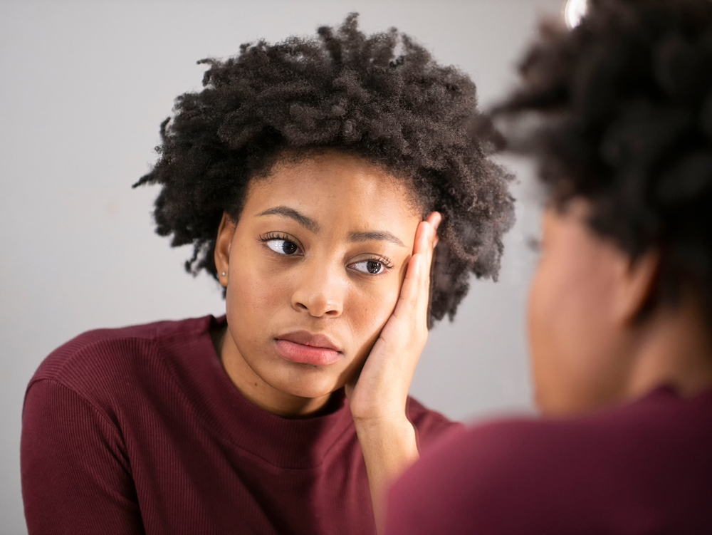 A woman looks at herself in the mirror and makes a depressed face.