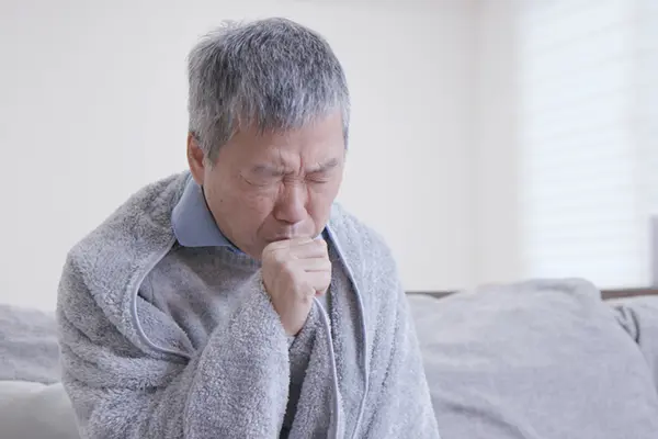 A man with a grey blanket over him is sitting on a bed or couch. He has his eyes closed and is coughing into his hand, which is shaped in a fist. 