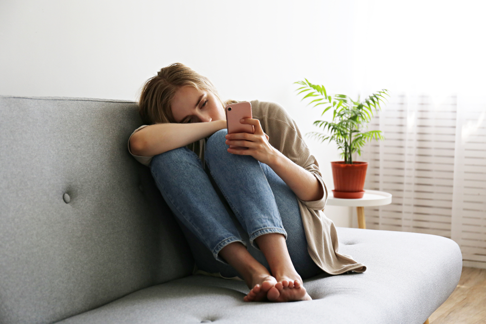 A woman sits curled up on the couch looking lonely and sad as she stares at her cell phone.