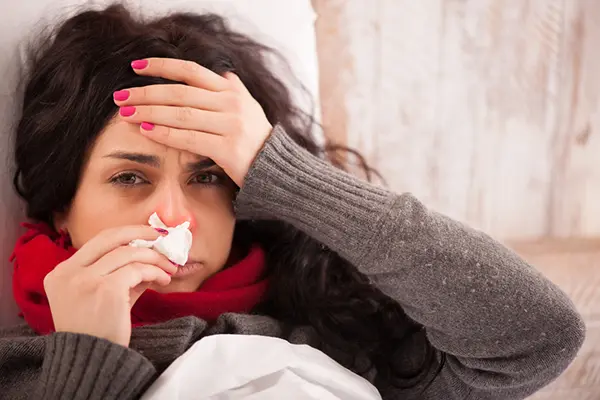 A brown-haired woman lies in a room with her head propped up on a pillow. Her eyes look tired and swollen, and she's holding a tissue to her nose. Her other hand is on her forehead. She's wearing a grey/brown sweater, and her nail polish is berry-colored. 