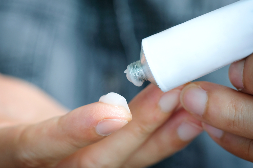 An adult squeezes a dollop of medicated cream out of a white tube onto their pointer finger.