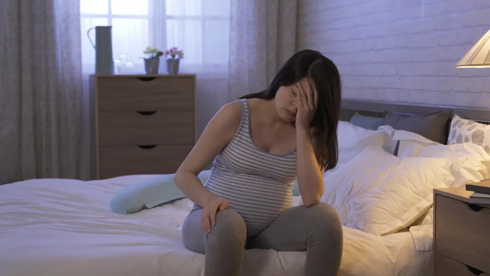 A pregnant woman sits on her bed with her head in her hand.