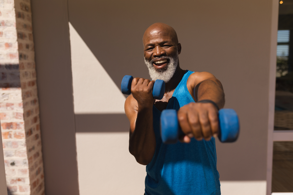A smiling man with a white beard and wearing a blue tank top. He has weights in his hands and is holding one out toward the camera. 