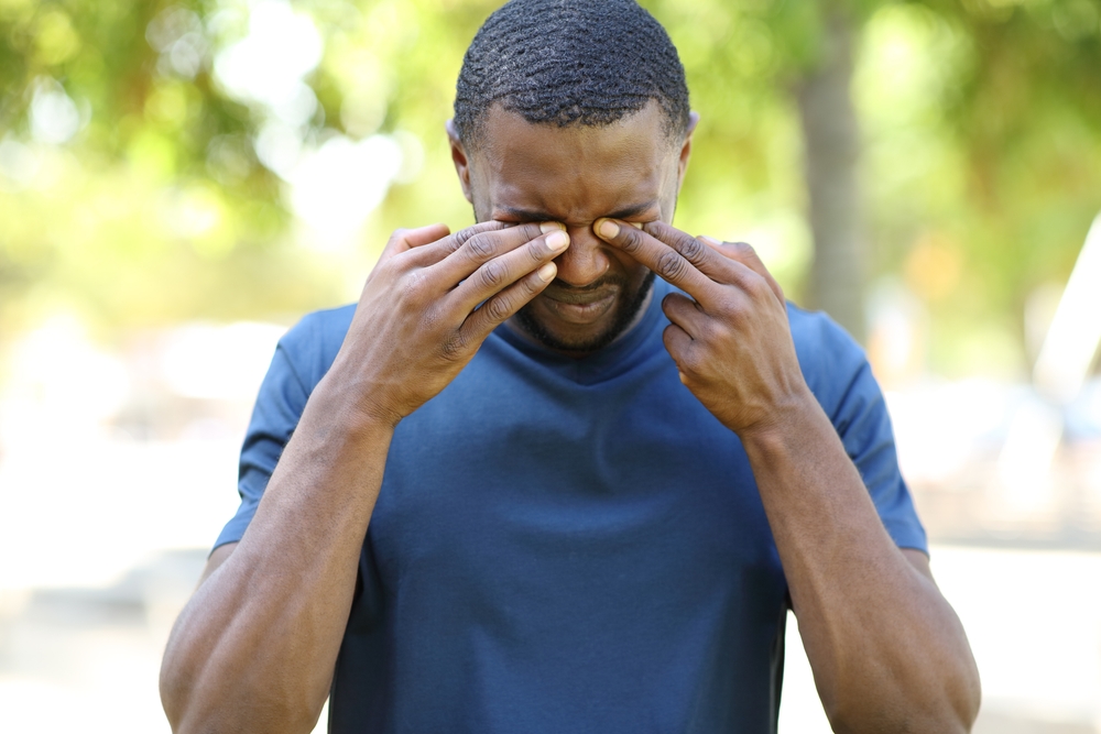 A man in a blue t-shirt stands outside while rubbing his eyes. 