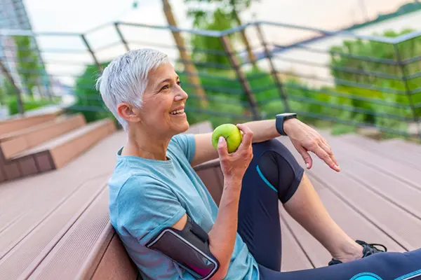 A fit-looking woman with cropped grey hair and a blue workout outfit is sitting outside on a set of stairs. She is holding an apple and smiling to someone or something off the camera. 