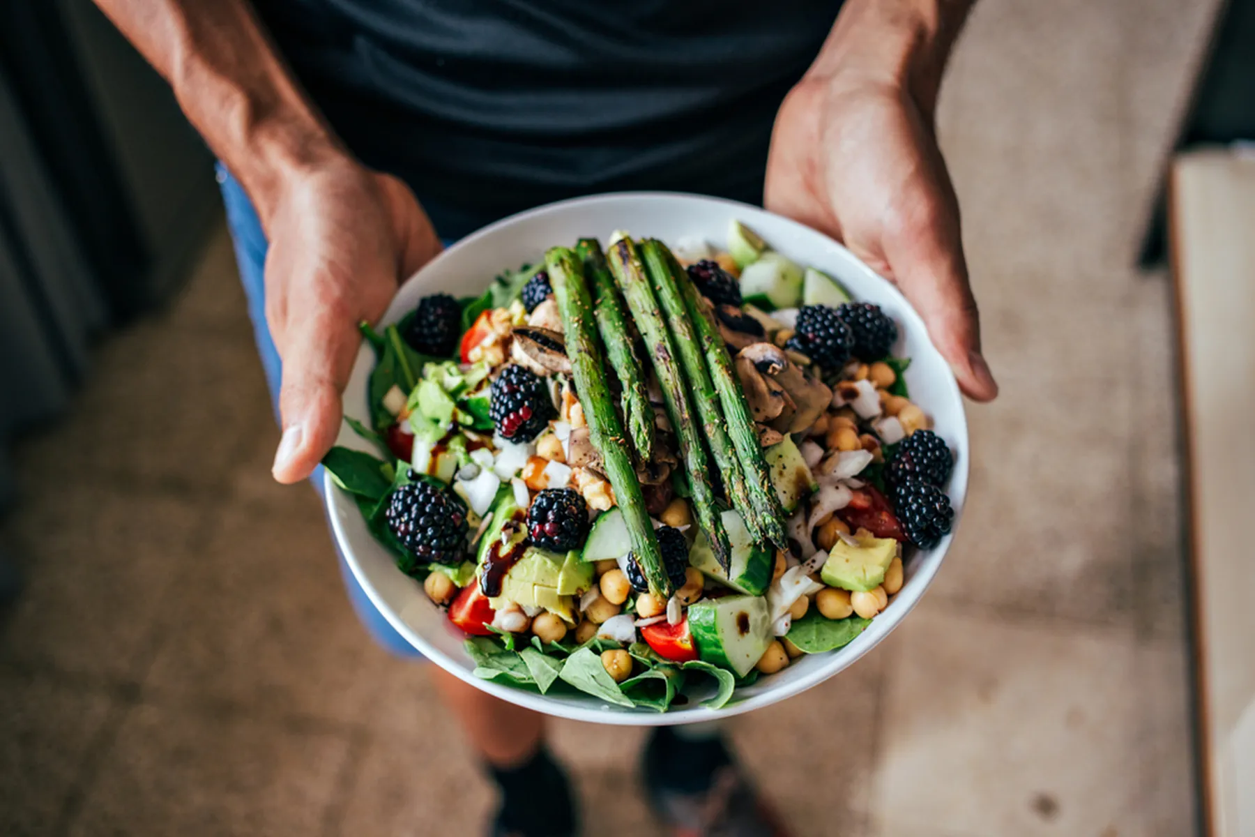 A man hands holds a salad with fruits, vegetables, and legumes such as asparagus, blackberries, cucumbers, and beans.