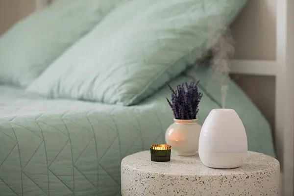 A diffuser sits next to a bed and diffuses essential oils into the air.