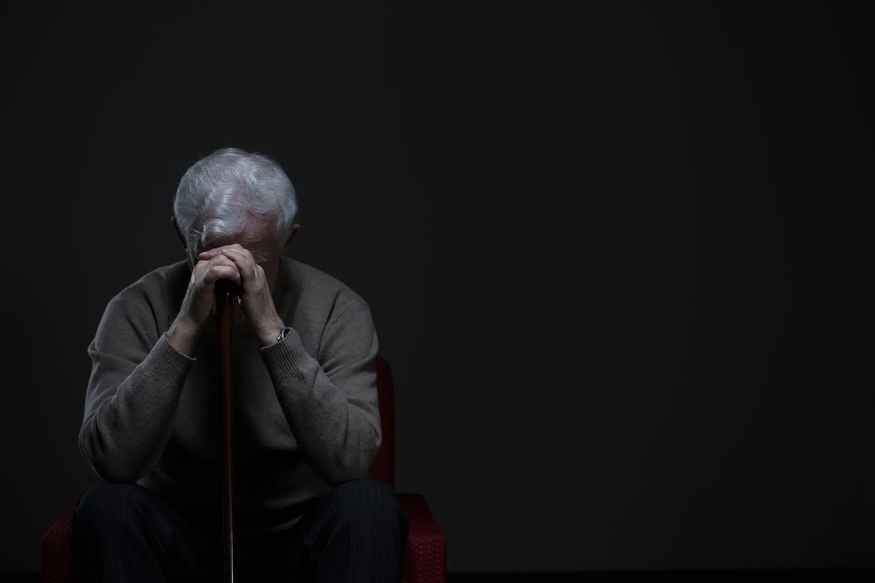 An older man holds his head to his cane as he sits in a dark room.