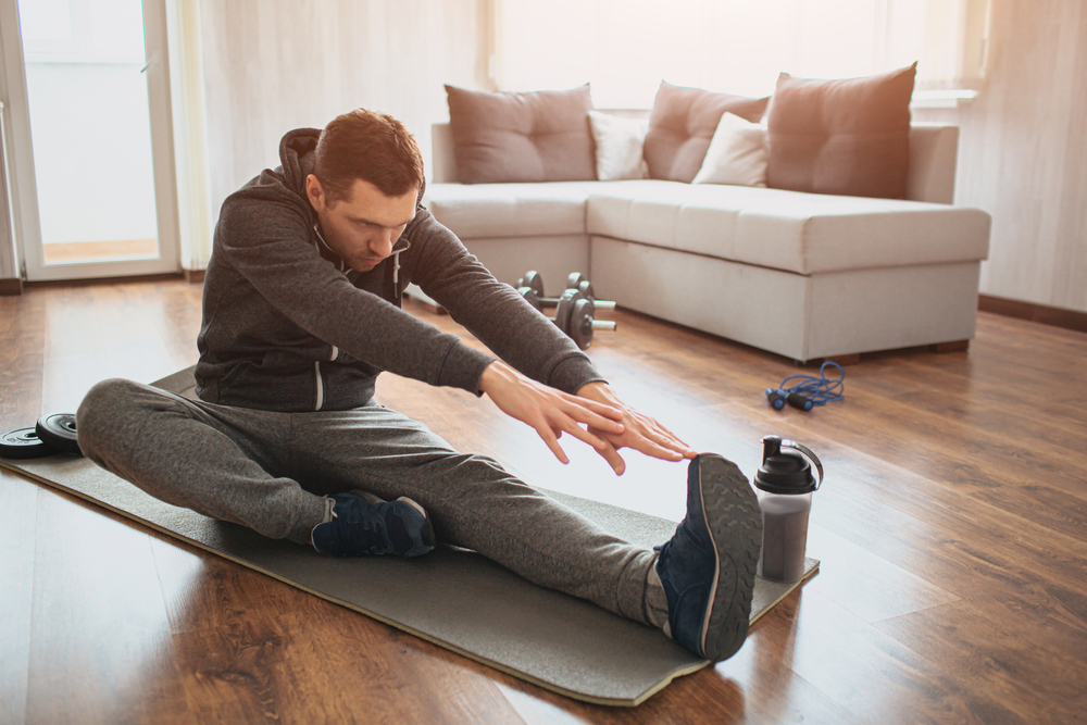 Man on a yoga mat on a hardwood floor. He has one leg in front of him and is stretching down toward it. There is a water bottle at his feet; behind him is an off-white couch with earth-colored cushions. 