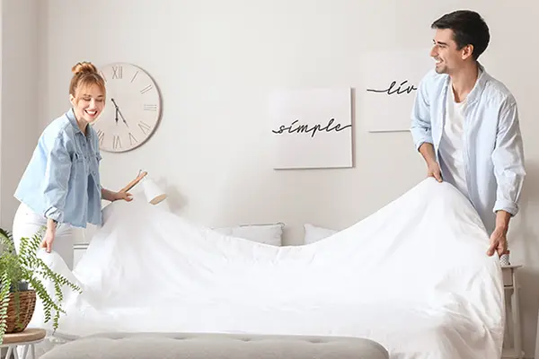 A couple is smiling while making their bed together.