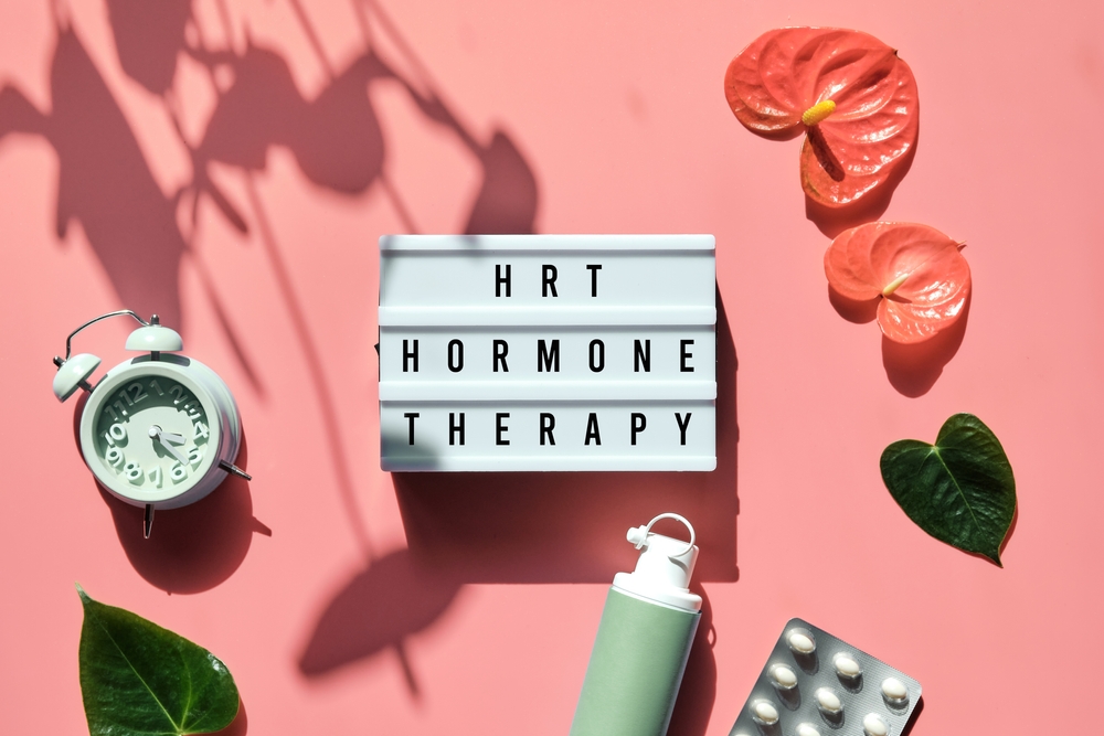 Sign reading: HRT Hormone Therapy rested on a pink background surrounded by flowers, a clock, and hormone gels and pills.
