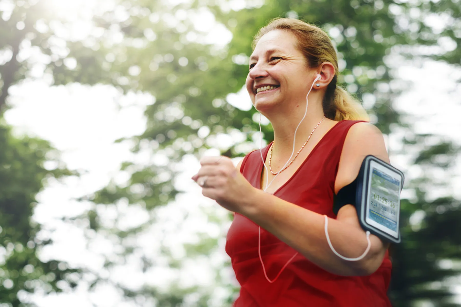 A woman in a red workout tank, an ipod on her arm, and earbuds in her ears smiles as she jogs or speed walks outside. 