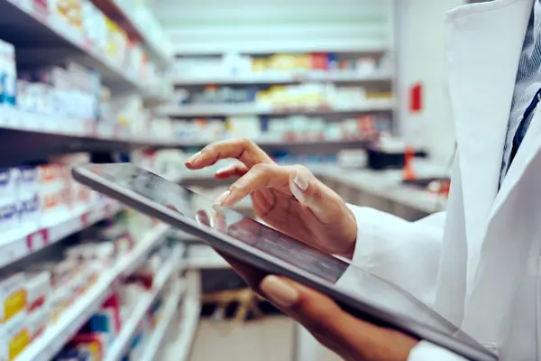 A pharmacist refers to an ipad while standing in front of shelves of medications. 