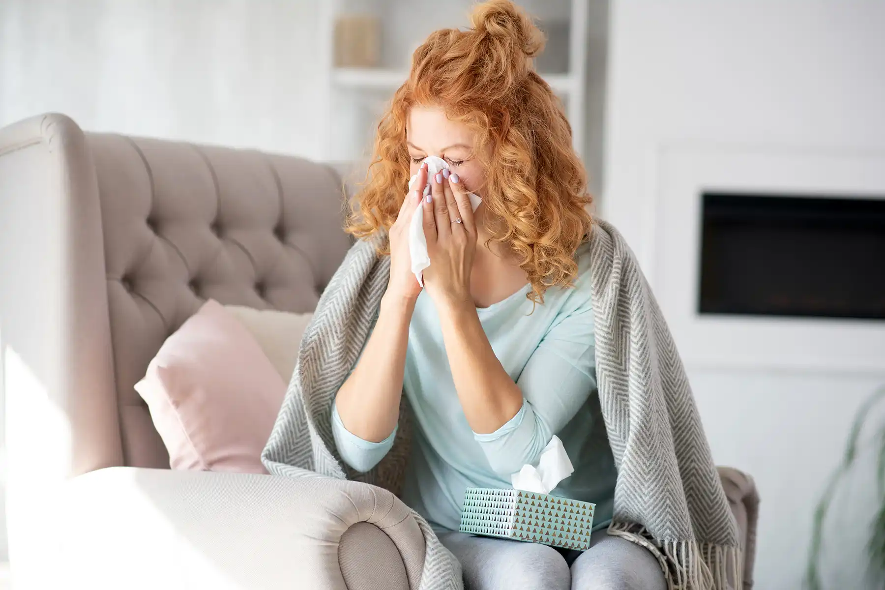 Women sitting on her couch covered in a blanket and blowing her nose.