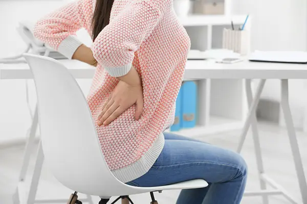 A young woman sitting in a chair at a desk. Her back is arched and she has her hands on her back in discomfort. 