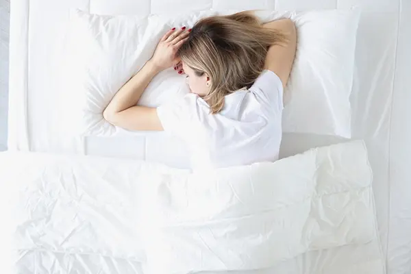 A woman in a white shirt sleeping on her stomach in a bed with white linens.