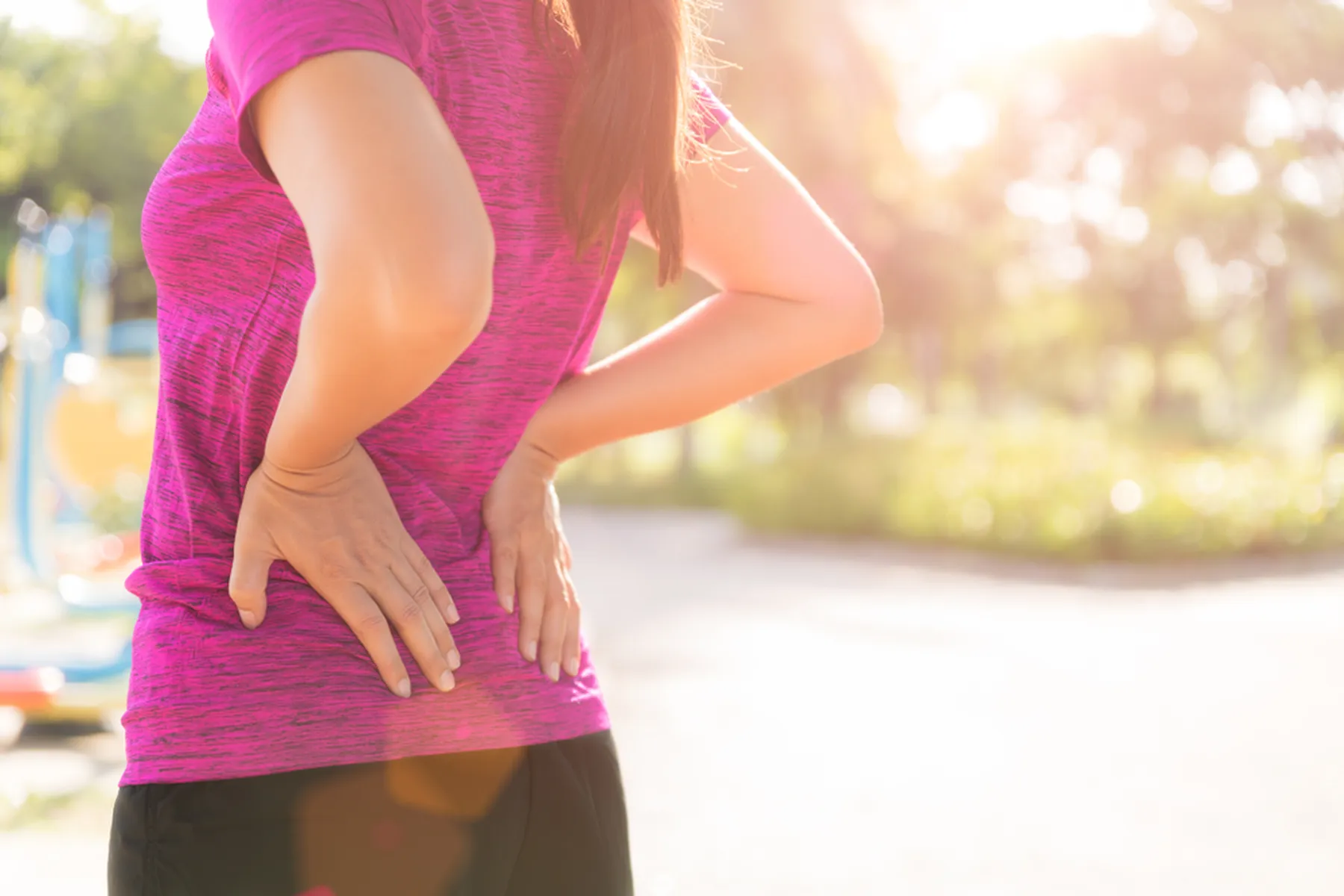 A woman in a dark pink t-shirt is holding both hands on her lower back in discomfort. 