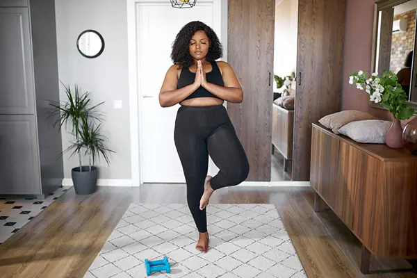 A woman stands in her living room in a yoga pose (tree pose) with her eyes closed.