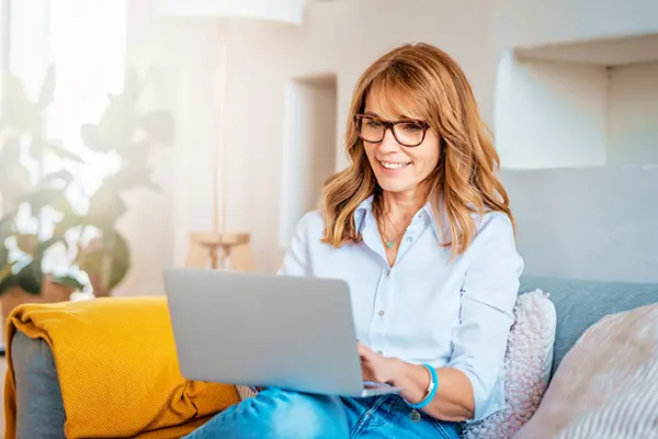 A woman in a light blue shirt and jeans sits on a couch as she looks at her laptop screen and smiles