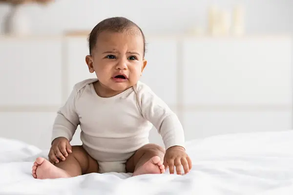 A baby in a white shirt and diaper sitting on the bed. The baby looks like they're starting to cry. 