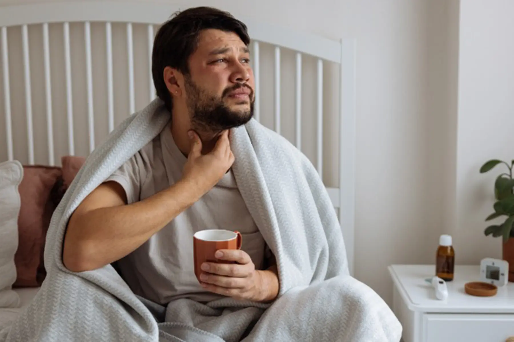 Man sitting in bed, wincing at his sore throat, while holding a cup of tea. 