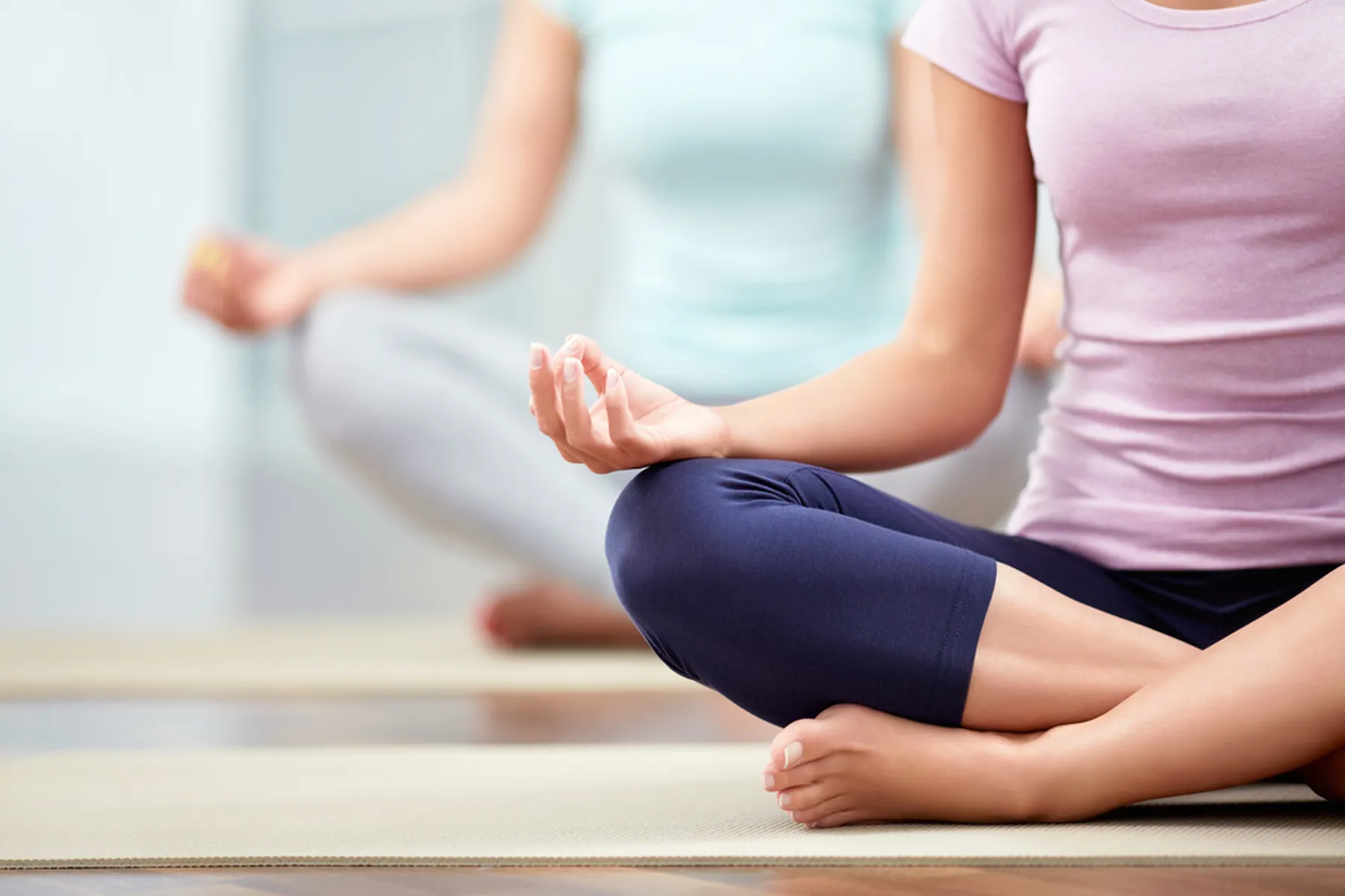 Two women are sitting in a meditative yoga pose with their palms resting on their knees, facing upwards. 