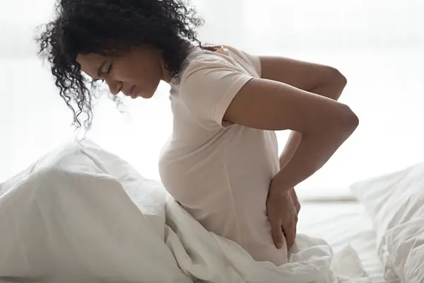 A woman sits up in bed with her hand on her lower back. She appears to be in discomfort. 
