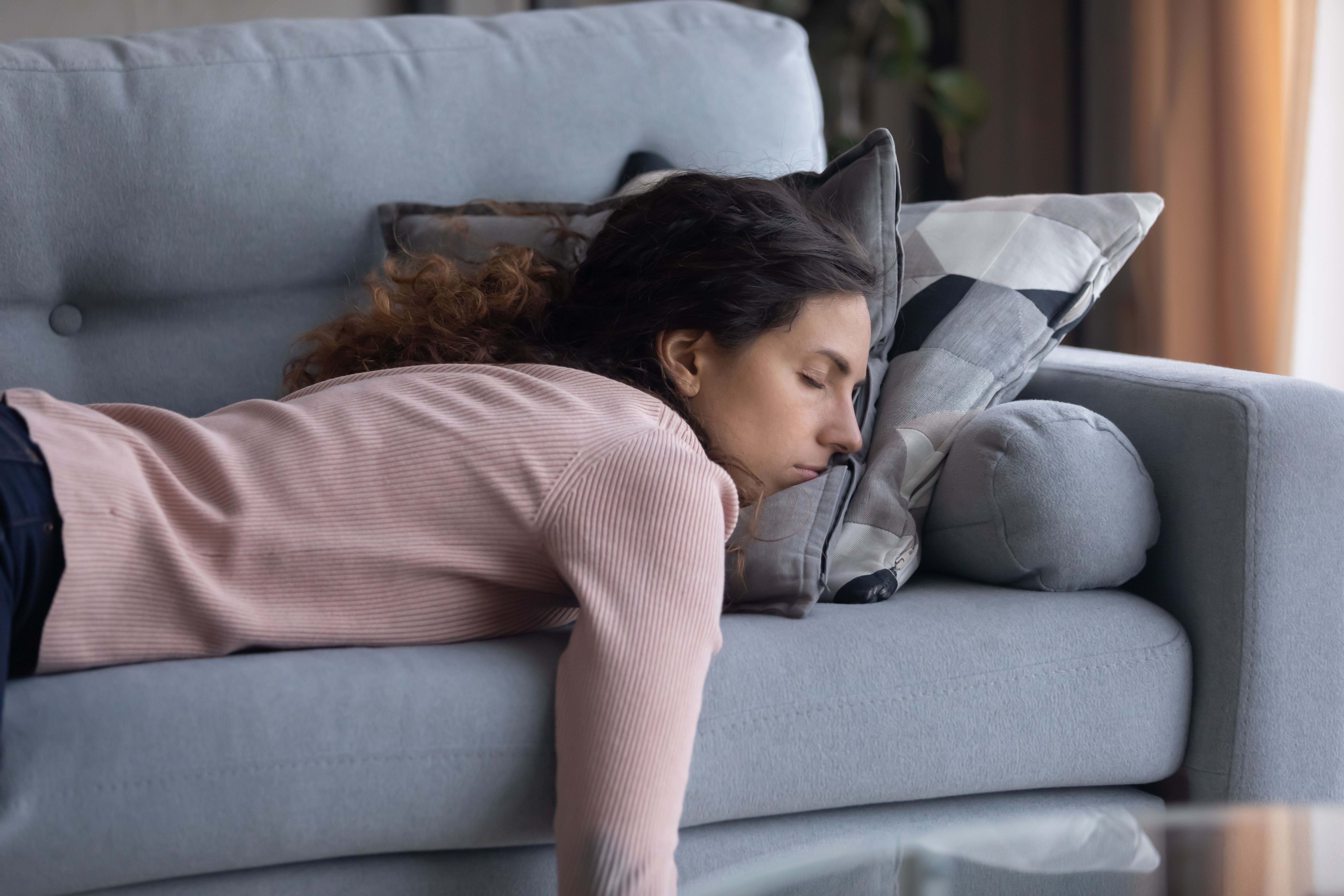 An exhausted woman sleeps on a grey couch. Her head is on two pillows as she sleeps on her stomach and her right arm hangs off the couch. 