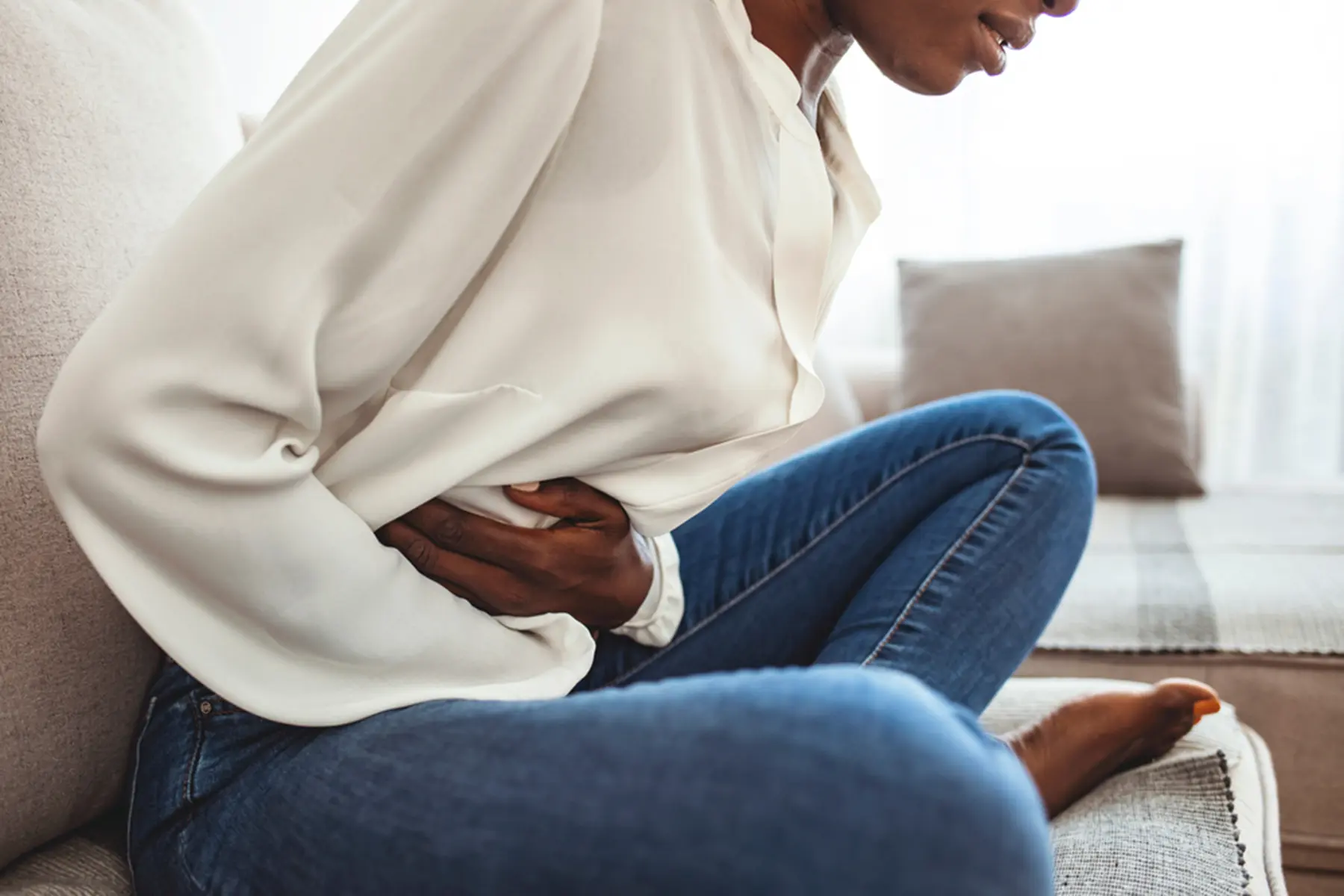 A woman sitting on a couch is doubled over in pain, clutching her stomach.
