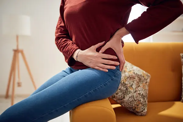 Woman sitting on the arm of a couch and holding her hands to her hip.
