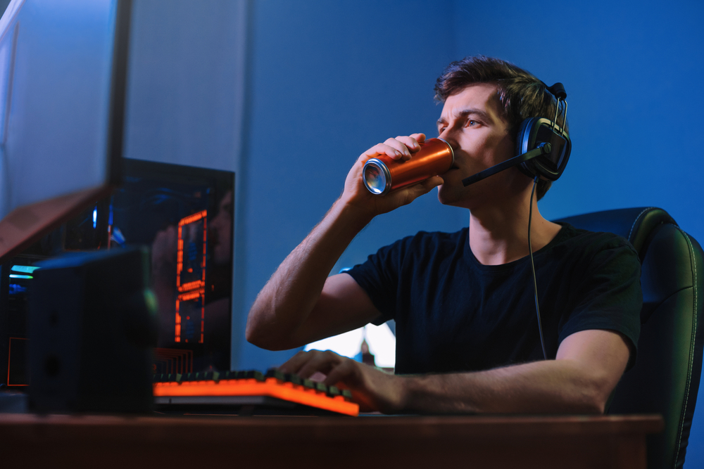 A man sits at his computer in a dark room and drinks an energy drink.
