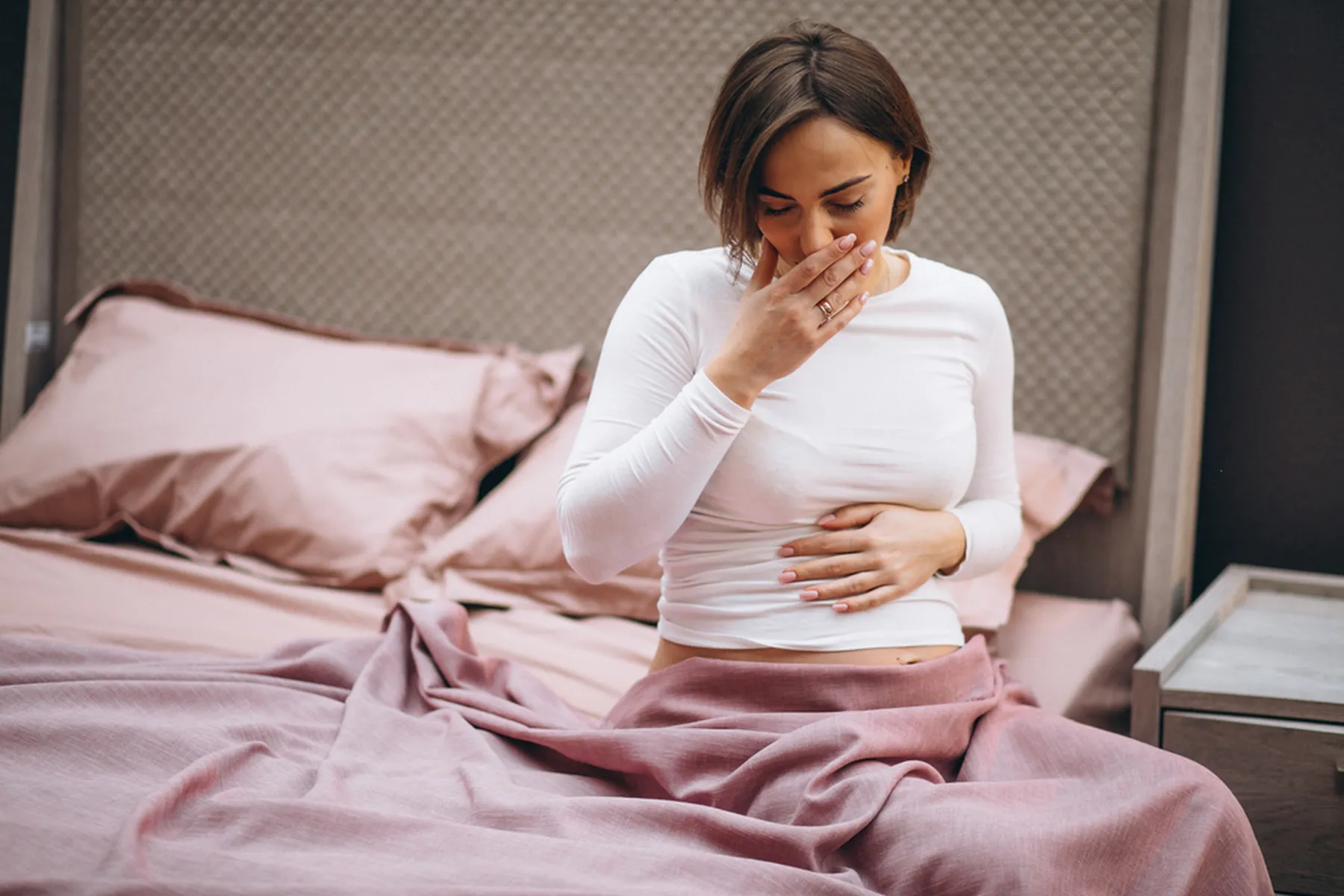A woman in her first trimester of pregnancy sits on her bed and clutches her stomach with one hand and mouth with the other.