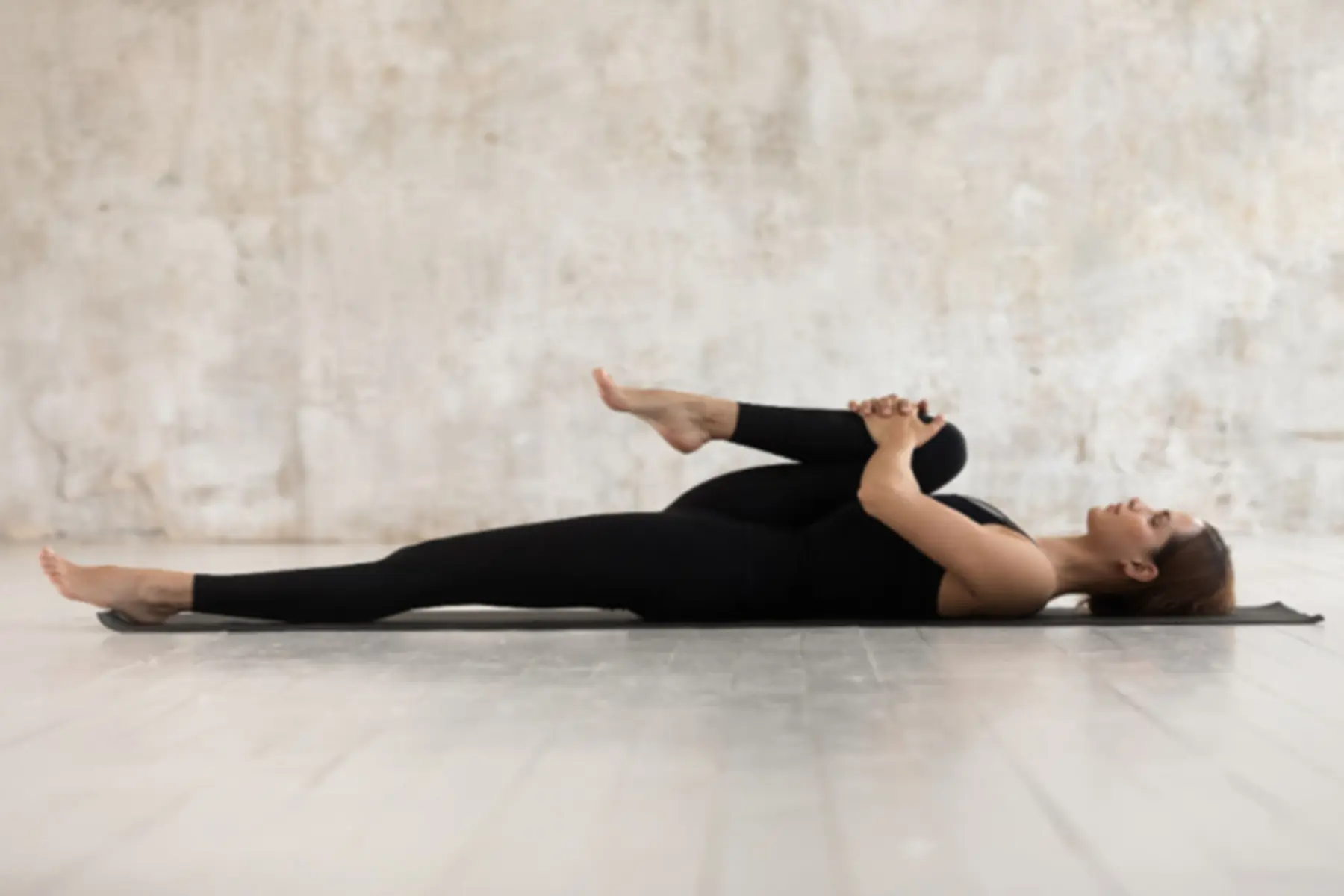 A woman lays on a yoga mat and stretches her knee to her chest.