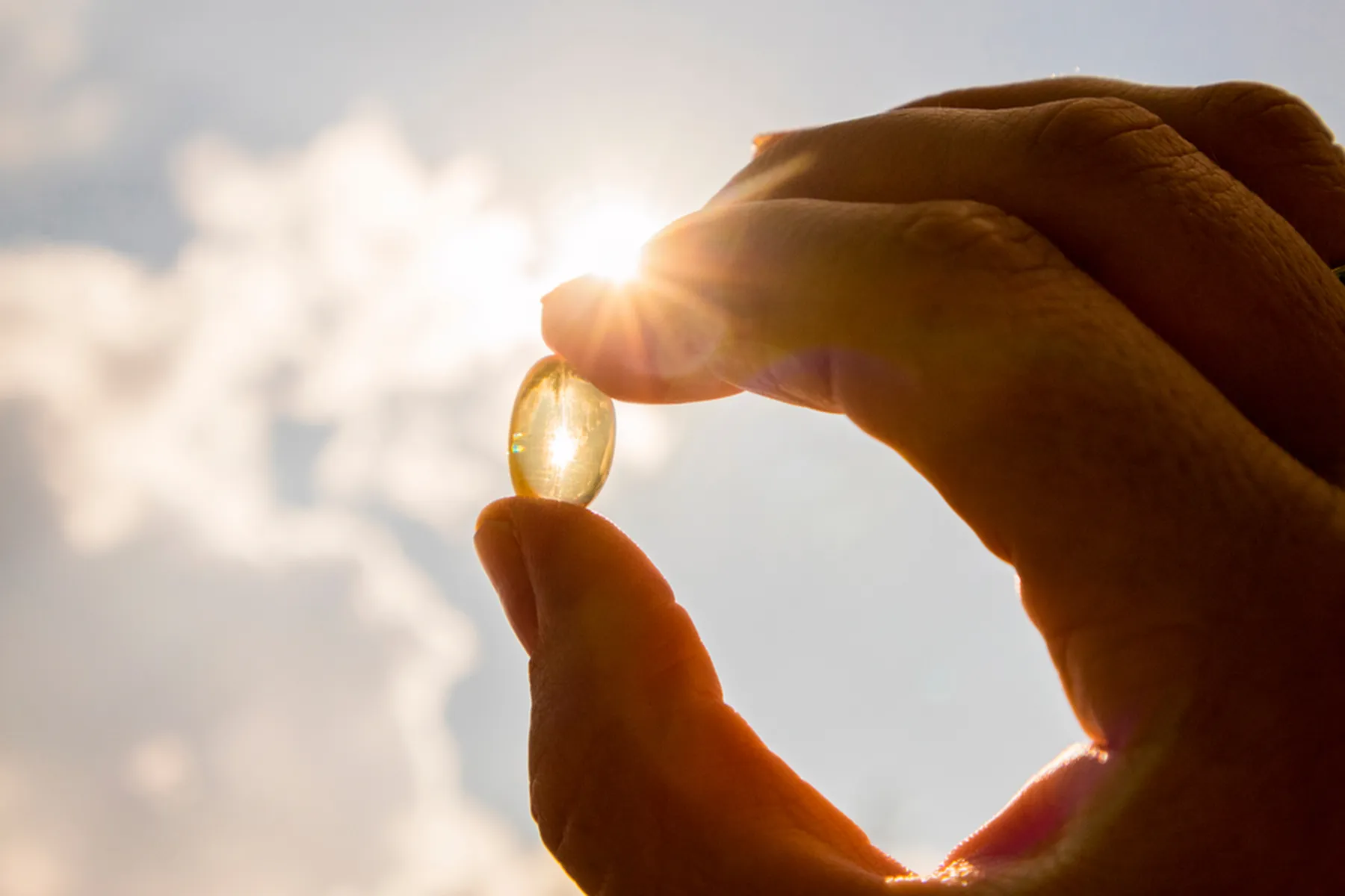 A vitamin d supplement capsule is held up to the sky, with sunlight shining through it.