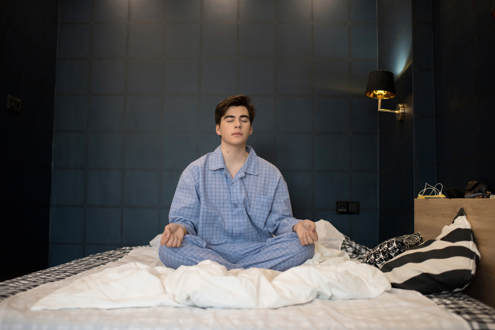 A man sits on his bed in his pajamas and meditates.